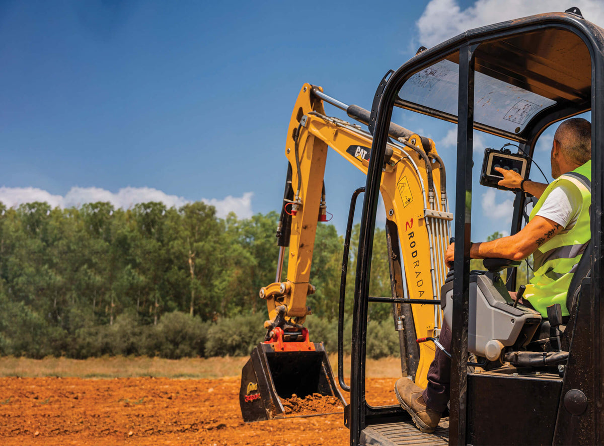 A yellow excavator with a visible ROKA brand mark is being operated by a worker inside a black cab in an outdoor construction setting with red soil and trees under a blue sky.